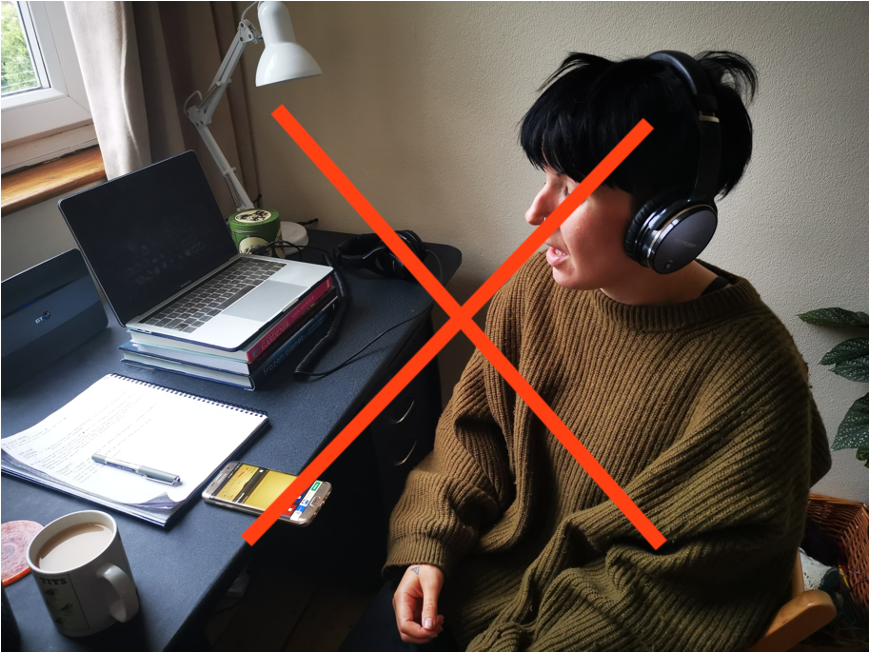 Photo of someone sitting at a desk with headphones on. They are speaking into their phone, which is lying on the desk. There is a big red cross across the photo. 