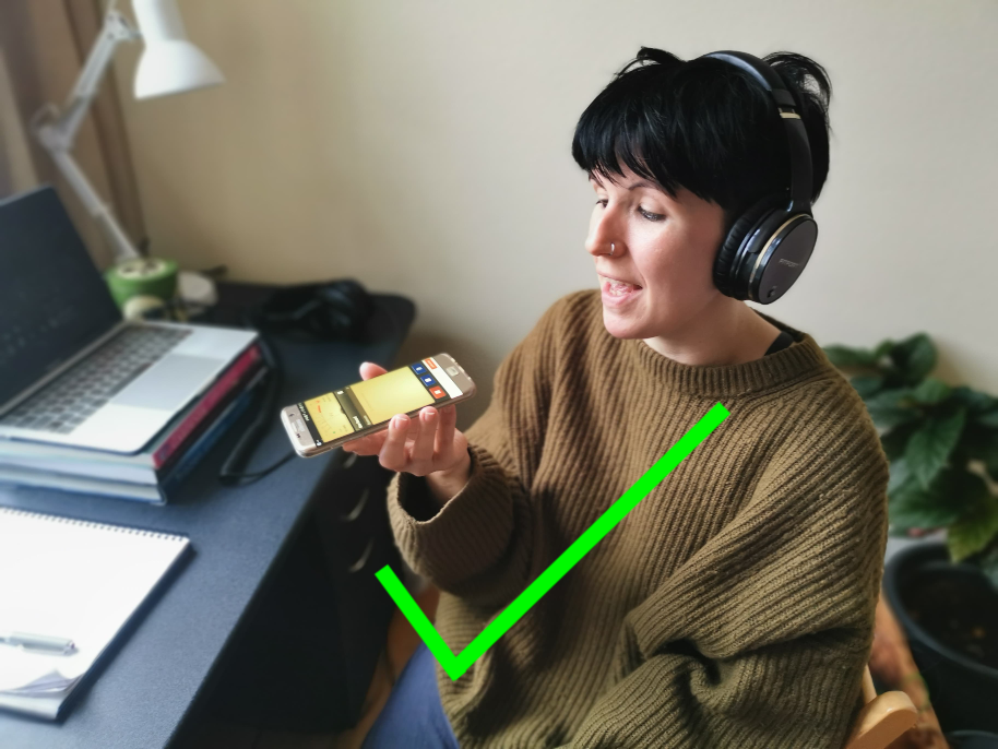 Photo of someone sitting at a desk with headphones on. They are speaking into their phone, which they are holding with the microphone about 15cm from their mouth. There is a big green tick across the photo. 